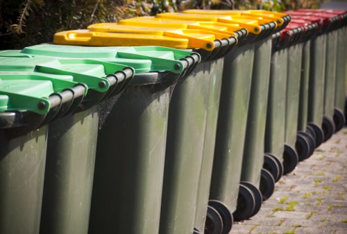 A row of multiple large plastic waste bins lined up along a paved surface, each with color-coded lids in green, yellow, and red. The bins are designed for rubbish collection and are placed closely together, with black wheels visible at the bottom. The background features blurred greenery, indicating an outdoor environment, possibly near a commercial or residential area in Blackfriars. The image is associated with commercial waste removal services, highlighting organized rubbish storage solutions managed by companies like Commercial Waste Removal Blackfriars. The lighting suggests daylight, casting soft shadows on the ground and the bins, emphasizing their smooth, slightly reflective surfaces and the vibrant color of the lids for easy identification.