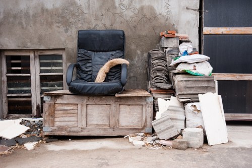 The image shows a scene of waste accumulation outdoors, featuring a black office chair with a worn beige cushion placed on a weathered wooden chest. To the right of the chair, there is a stack of building materials including concrete blocks, bricks, and cement boards, some of which are broken or chipped. The objects are situated on a paved surface next to a rough, stained concrete wall with a small ventilation grate and a partially visible gate or door in the background. Debris and small pieces of rubble are scattered at the base of the stack, indicating discarded or dropped construction debris. This scene likely depicts a rubbish removal scenario in an urban or industrial area that a service such as Commercial Waste Removal Blackfriars might address, emphasizing the need for professional clearance of unwanted furniture and building waste from the environment.