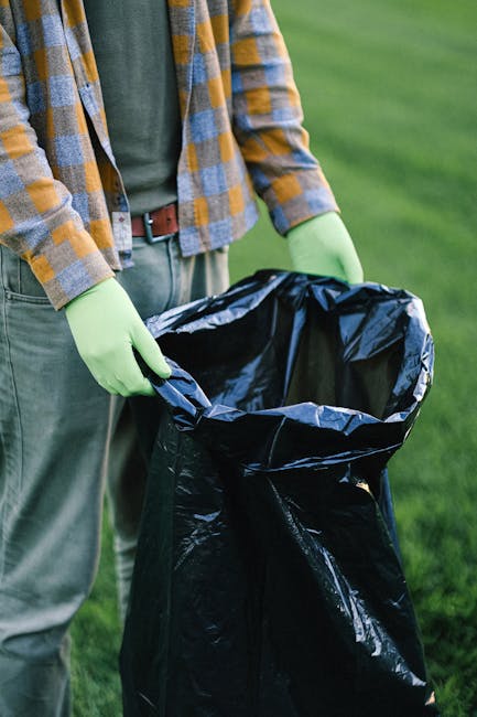 A waste collection worker dressed in a green uniform and helmet is standing on the side of a street near a commercial rubbish bin, which appears to be heavy-duty and made of metal, with its rear compa