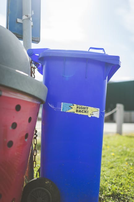 The image shows a large, upright blue plastic rubbish bin equipped with a lid, positioned outdoors on a grassy area near a pavement. The bin has a label with the words 'FIGHT BACK!' on its front, indi