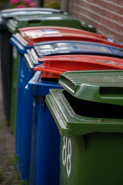 The image shows a large, upright blue plastic rubbish bin equipped with a lid, positioned outdoors on a grassy area near a pavement. The bin has a label with the words 'FIGHT BACK!' on its front, indi